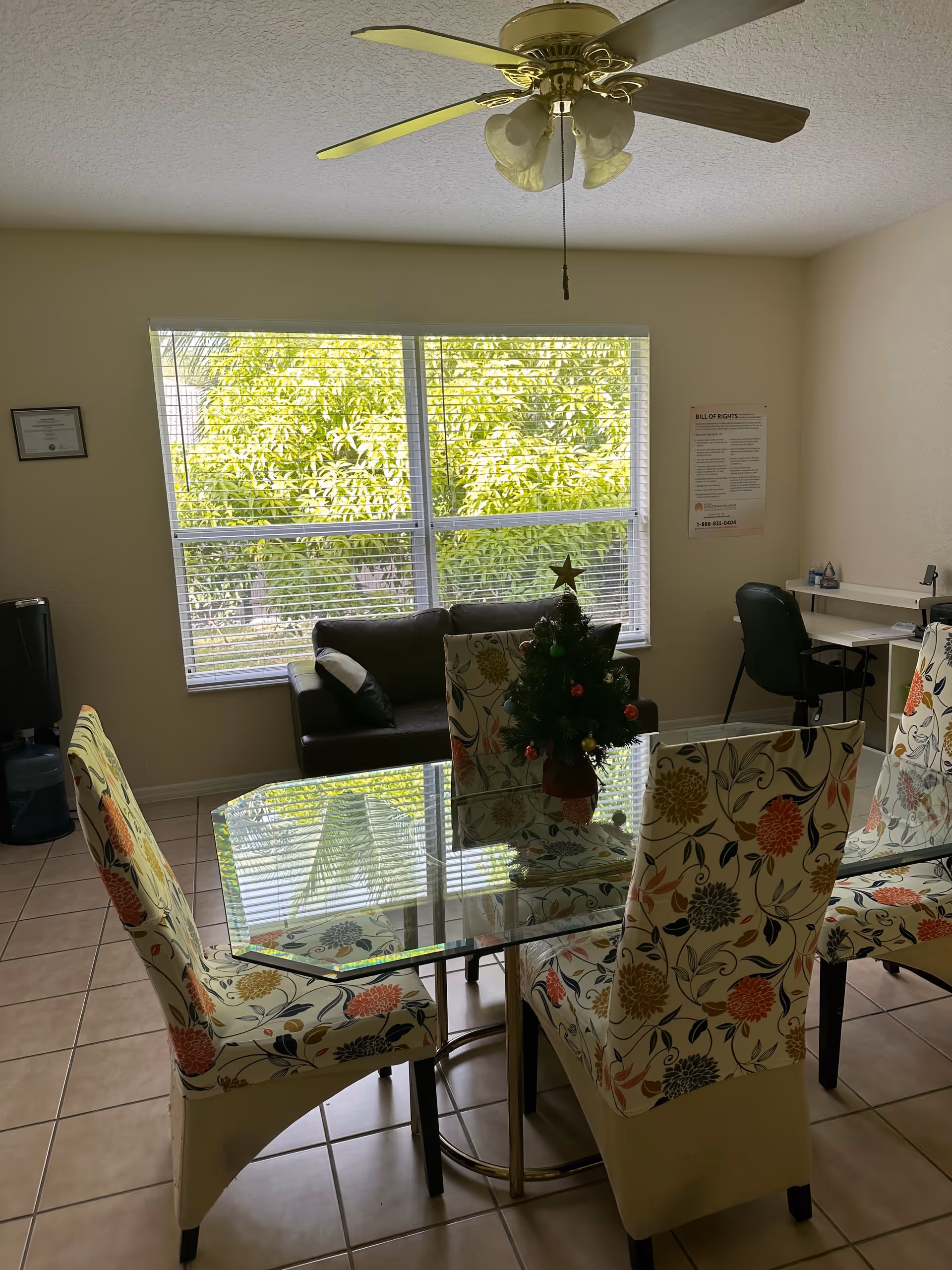 A bright room with a glass dining table surrounded by four floral-patterned chairs. A small decorated Christmas tree sits on the table. In the background, there is a brown couch in front of a large window with blinds, a ceiling fan with lights, a water dispenser, and a desk with a black office chair. The walls are light-colored and the floor is tiled.