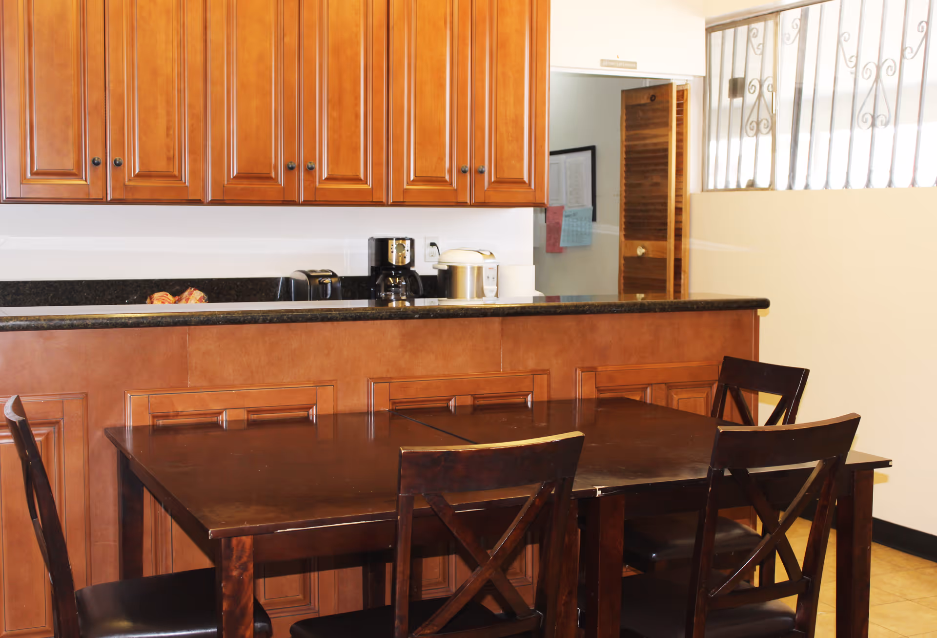 Interior view of a kitchen area with wooden cabinets, a black countertop, and a dark wooden dining table with four chairs. Appliances including a coffee maker, toaster, and rice cooker are on the counter. A door and a window with metal bars are visible in the background.