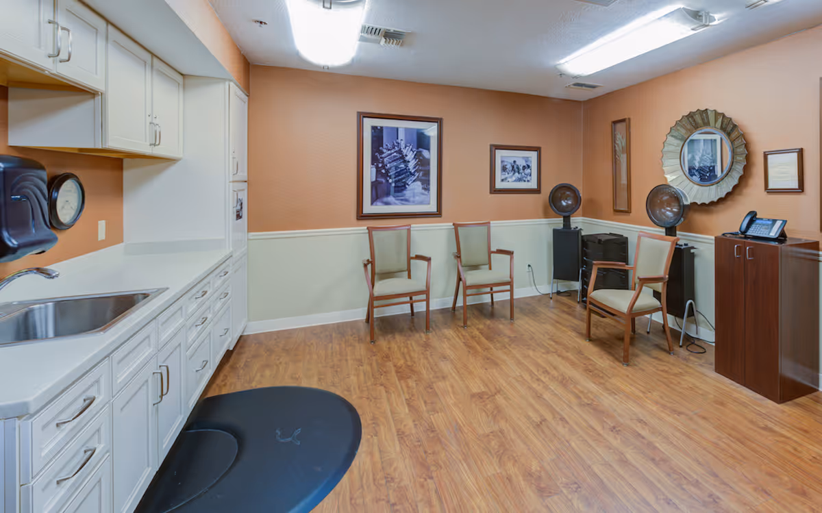 Interior room with wooden flooring and beige walls, featuring white cabinets and a sink on the left side. There are four wooden chairs with light upholstery arranged along the walls. The room also contains two vintage hair dryers, a round decorative mirror, framed pictures on the walls, and a small cabinet with a phone on top.