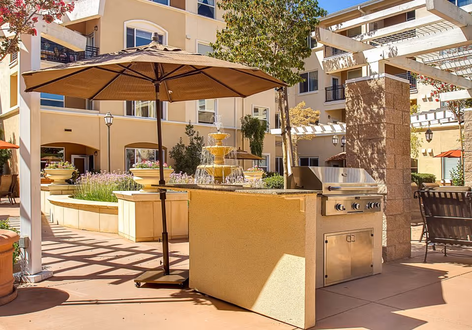 Outdoor patio area at Heritage Estate Senior Apartments featuring a built-in stainless steel grill, a large brown patio umbrella, a multi-tiered water fountain, potted plants, and seating areas with trees and a multi-story building in the background.