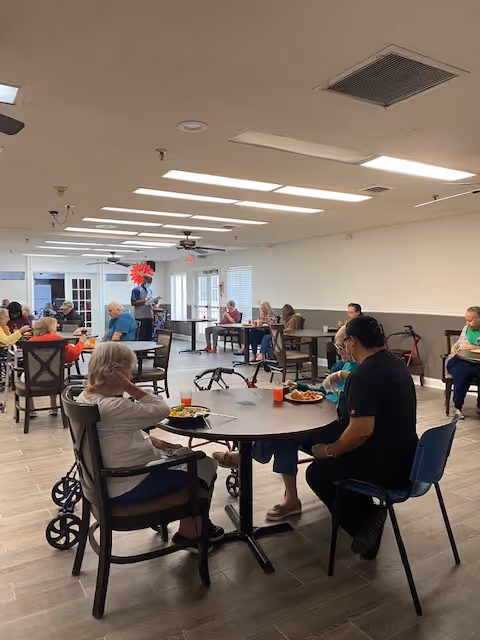 A dining room in a senior living facility with several elderly residents seated at round tables eating meals. The room has a neutral color scheme with beige walls and tiled floors. There are ceiling fans and fluorescent lights overhead, and some residents use walkers or wheelchairs.