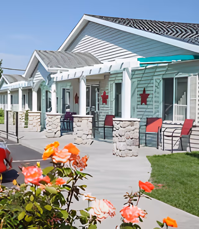 Front exterior of a single-story senior living building with a covered porch, stone columns, red chairs, and roses in the foreground.