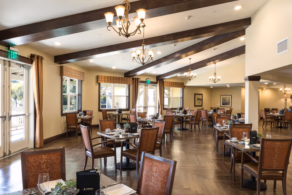 Spacious dining room with multiple set tables, wooden ceiling beams, chandeliers, and large windows.