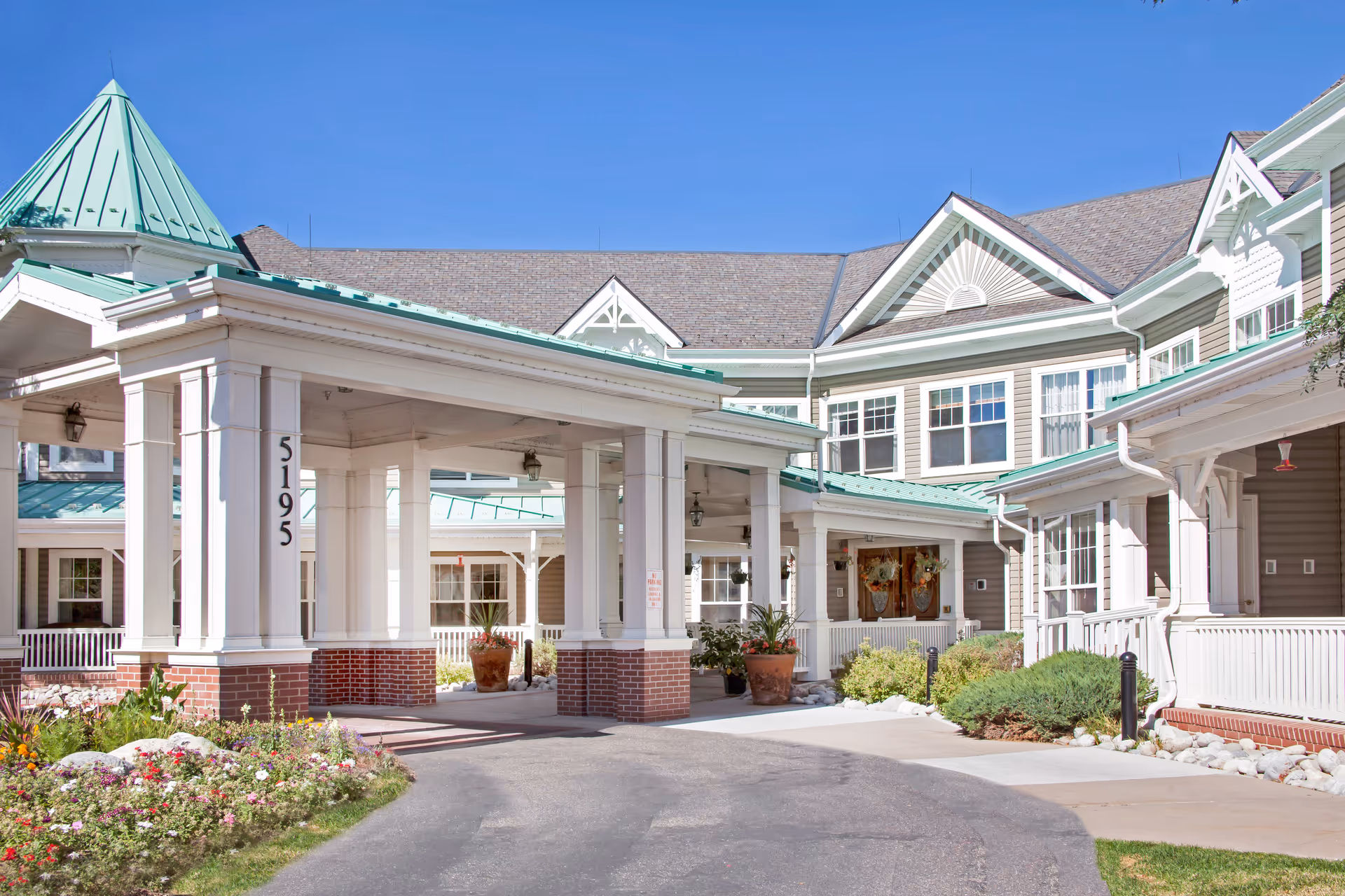 Front entrance and porte-cochere of a senior living facility with white columns, green roofs, and landscaped flower beds.