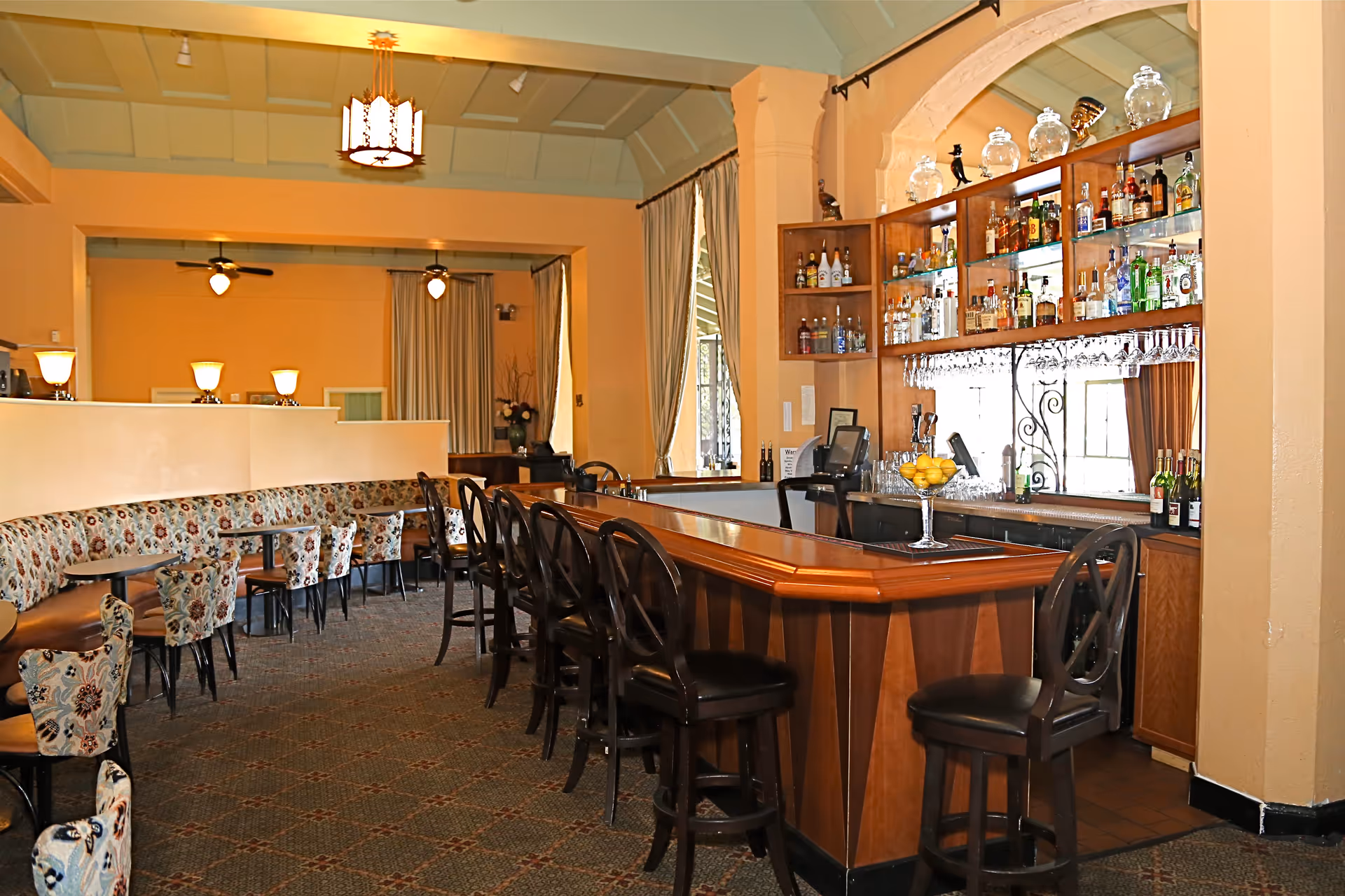 Interior view of a bar area with a wooden counter, several black bar stools, and shelves stocked with various bottles of liquor and glassware. To the left, there are tables with floral upholstered chairs and a long cushioned bench along the wall. The room has warm lighting, beige walls, and large windows with curtains.
