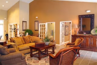 A cozy living room with a mustard yellow sofa, a brown armchair, and two wicker chairs around a wooden coffee table. The room has beige walls, a large mirror above a wooden cabinet, framed artwork, and a potted plant on the coffee table. There are open French doors leading to another room.