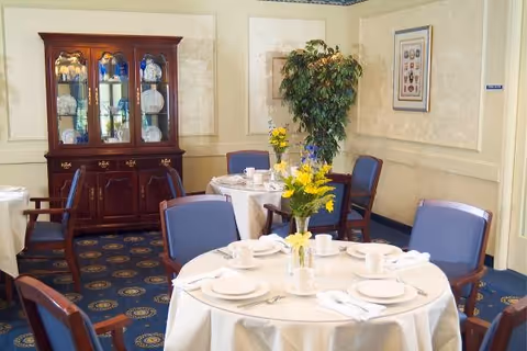 Round tables set for a meal with white tablecloths, blue upholstered chairs, floral centerpieces, and a china cabinet in a decorated dining room.