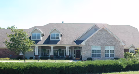 Front exterior view of a single-story senior living facility building with a combination of brick and stone facade, multiple windows, a covered entrance porch, and a well-maintained lawn with shrubs and a small tree.