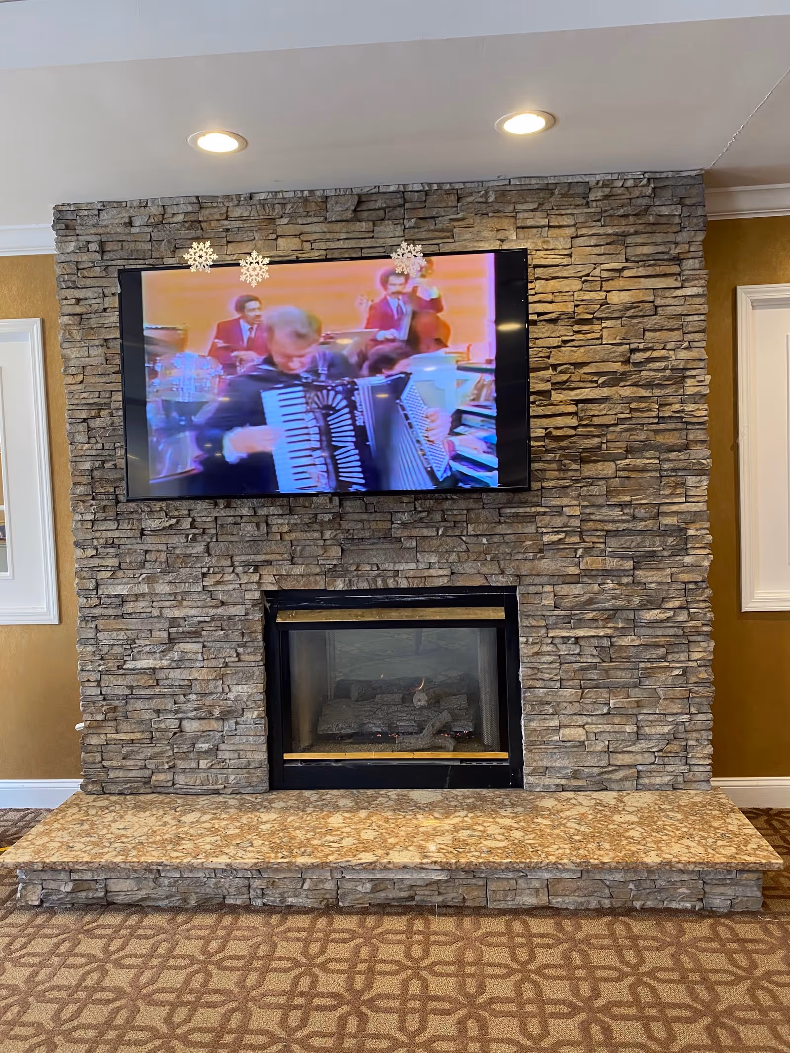 Stone fireplace with a mounted flat-screen TV showing musicians above a gas fireplace and a patterned carpeted floor.