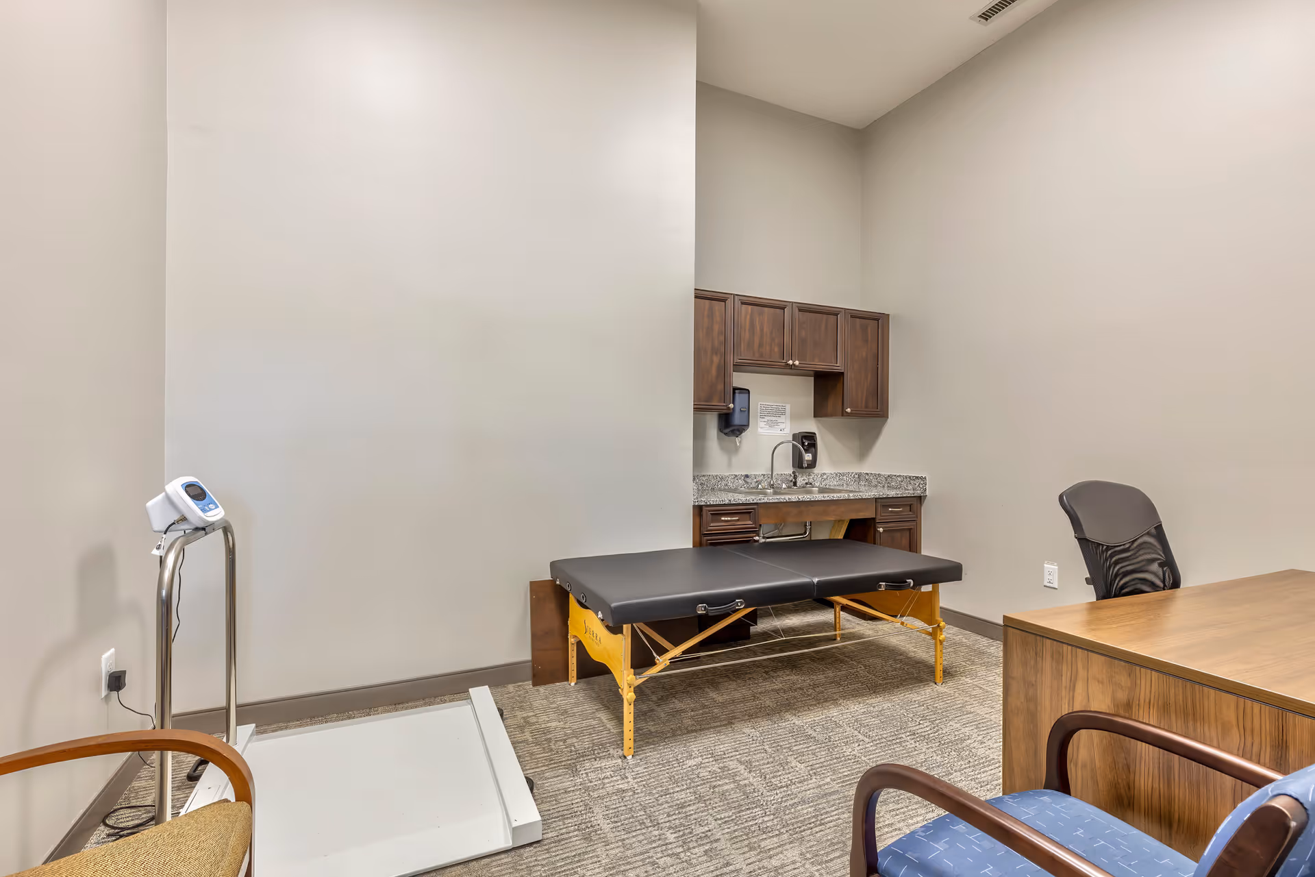 A small medical or therapy room with a black padded examination or massage table, a wooden desk with a black office chair, and two visitor chairs with wooden armrests. There is a white scale on the floor near the wall, and a countertop with a sink and wooden cabinets above and below it. The walls are painted light beige and the floor is carpeted.