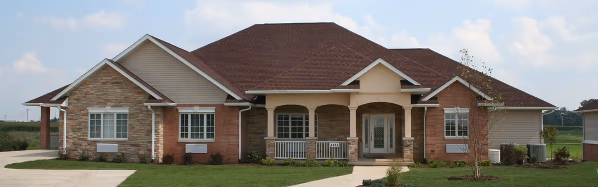 Front exterior view of a single-story senior living facility building with a combination of brick and stone facade, a covered porch with white railings, multiple windows, and a brown shingled roof under a partly cloudy sky.