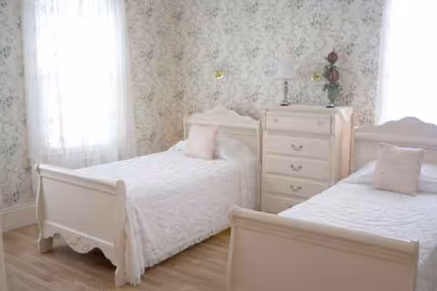 Two white twin beds with matching dresser and floral wallpaper in a bright bedroom.