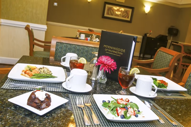 A dining table in a dining room set with plated meals, iced tea, a vase with a pink flower and a Morningside of Wilmington menu.