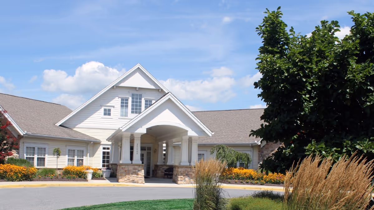 Front exterior view of Somerford House Frederick, a single-story building with a peaked roof and a covered entrance supported by columns. The building is surrounded by well-maintained landscaping including bushes, flowers, and trees under a partly cloudy blue sky.