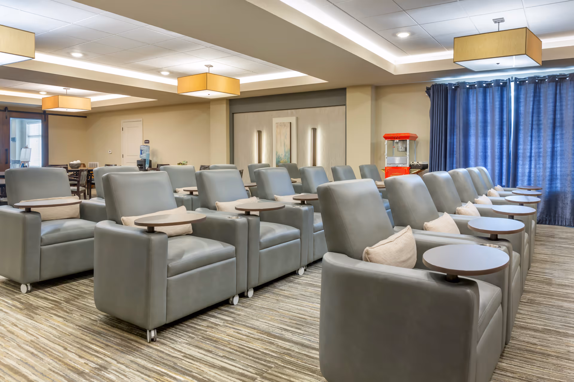 A spacious lounge area with multiple rows of gray reclining chairs, each equipped with a small round wooden table and a beige pillow. The room has a modern design with beige walls, a striped carpet, blue curtains covering the windows, and rectangular ceiling lights. In the background, there is a popcorn machine and a water dispenser near a wooden door.