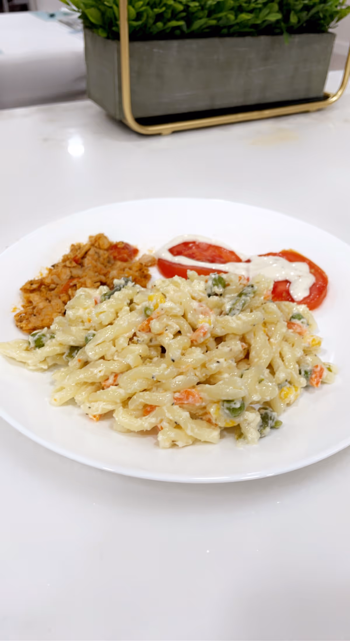 Plate of creamy spiral pasta salad with mixed vegetables, tomato slices with dressing, and a small side on a white table with a potted plant centerpiece.