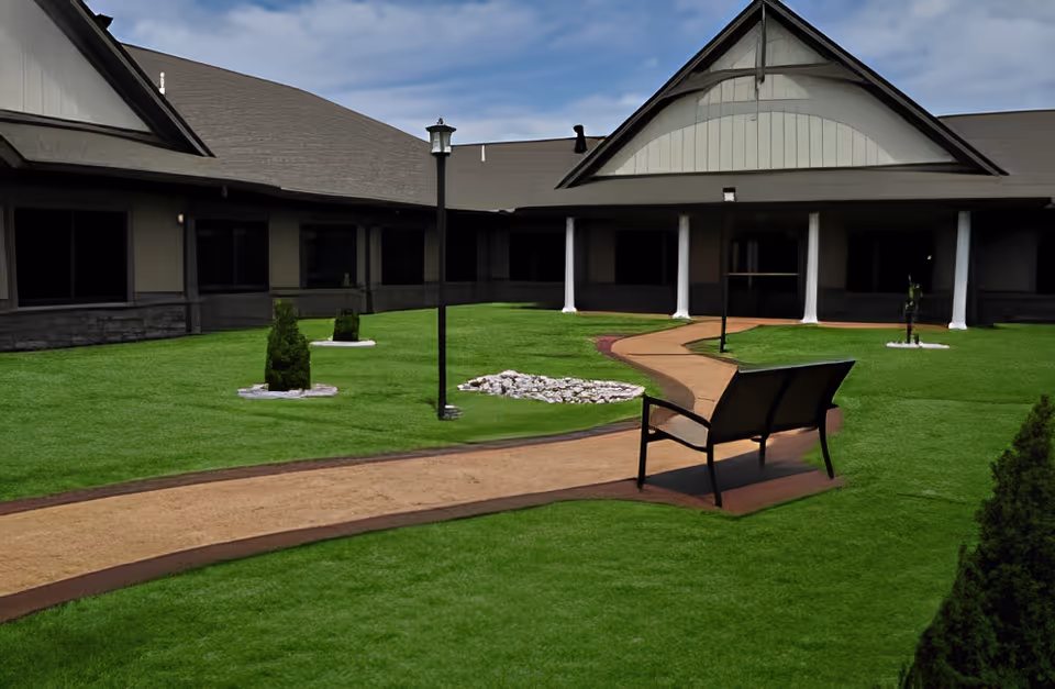A grassy courtyard with a winding walking path, a bench, small shrubs and a surrounding single-story building under a partly cloudy sky.