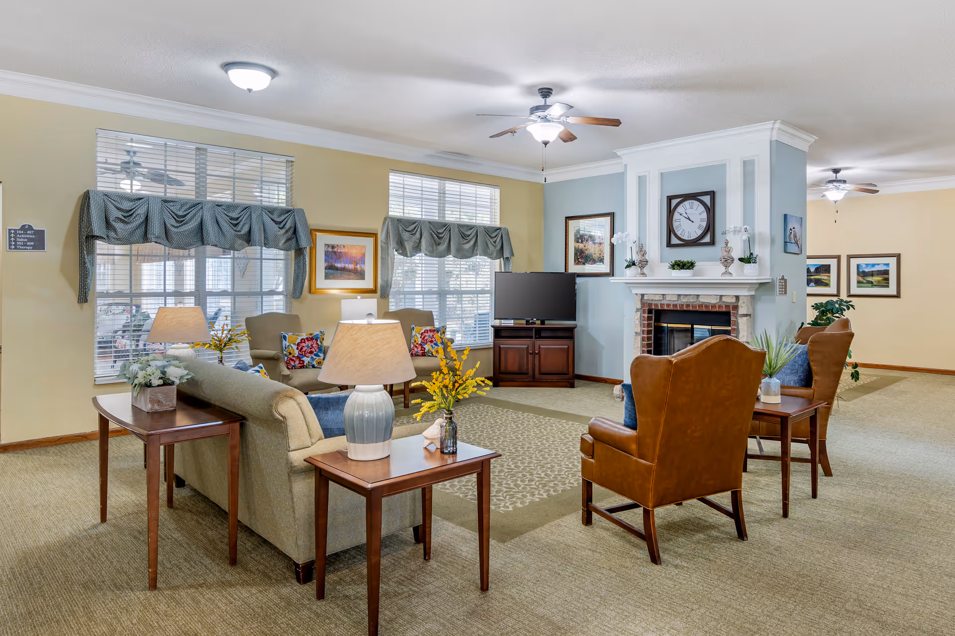 A cozy living room area in a senior living facility featuring a beige sofa with colorful pillows, two brown leather armchairs, wooden side tables with lamps and flower arrangements, a fireplace with a clock and decorative items on the mantel, a flat-screen TV on a wooden stand, large windows with green valances, ceiling fans, and framed artwork on the walls.