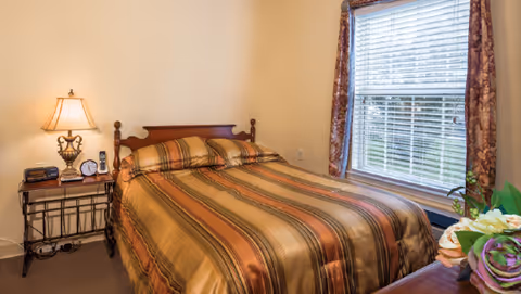 A bedroom with a double bed covered in a striped brown and orange bedspread. There is a bedside table with a lamp, clock, and phone on the left side of the bed. A window with floral curtains lets in natural light on the right side of the room. A small bouquet of flowers is visible on a piece of furniture in the foreground.