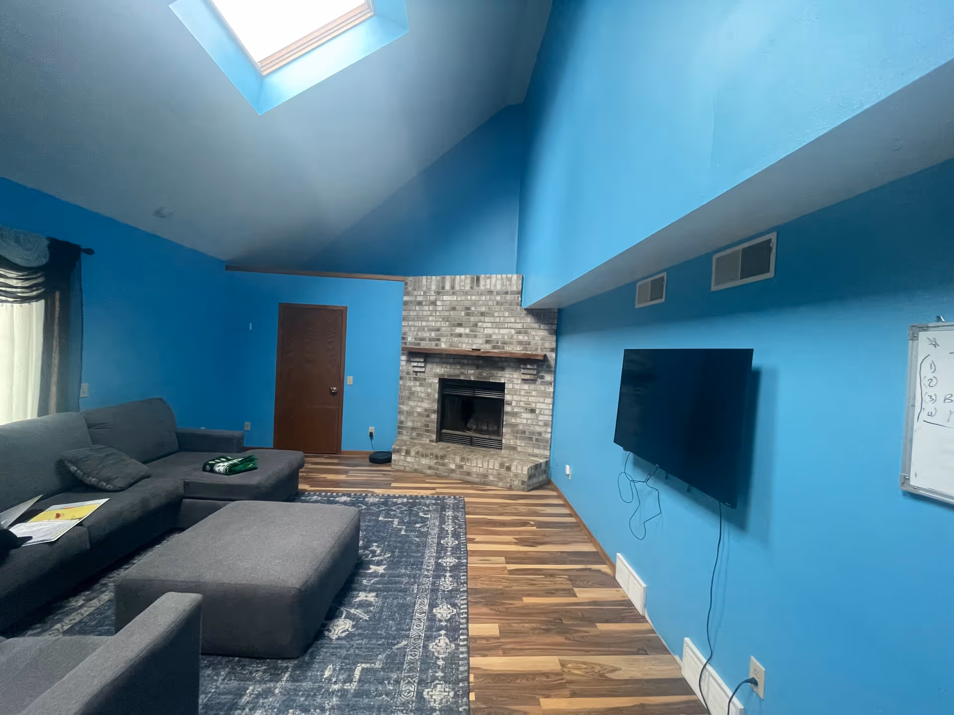 Interior of a living room with bright blue walls, a vaulted ceiling with a skylight, a gray sectional sofa with a matching ottoman, a patterned area rug, a brick fireplace, a wall-mounted flat screen TV, and a whiteboard on the wall.