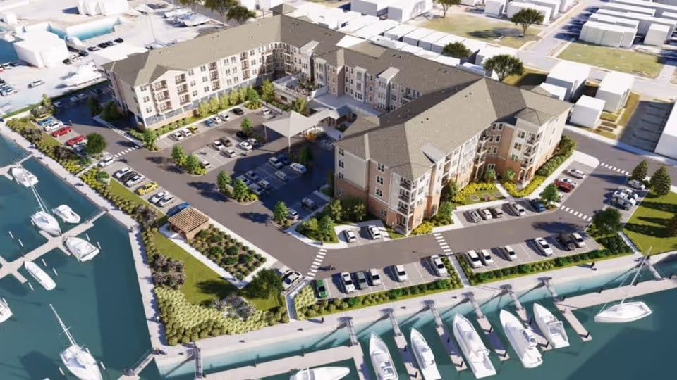 Aerial view of Acclaim At East Beach, a large senior living facility building with multiple floors surrounded by parking lots, landscaped greenery, and a marina with several docked boats along the waterfront.