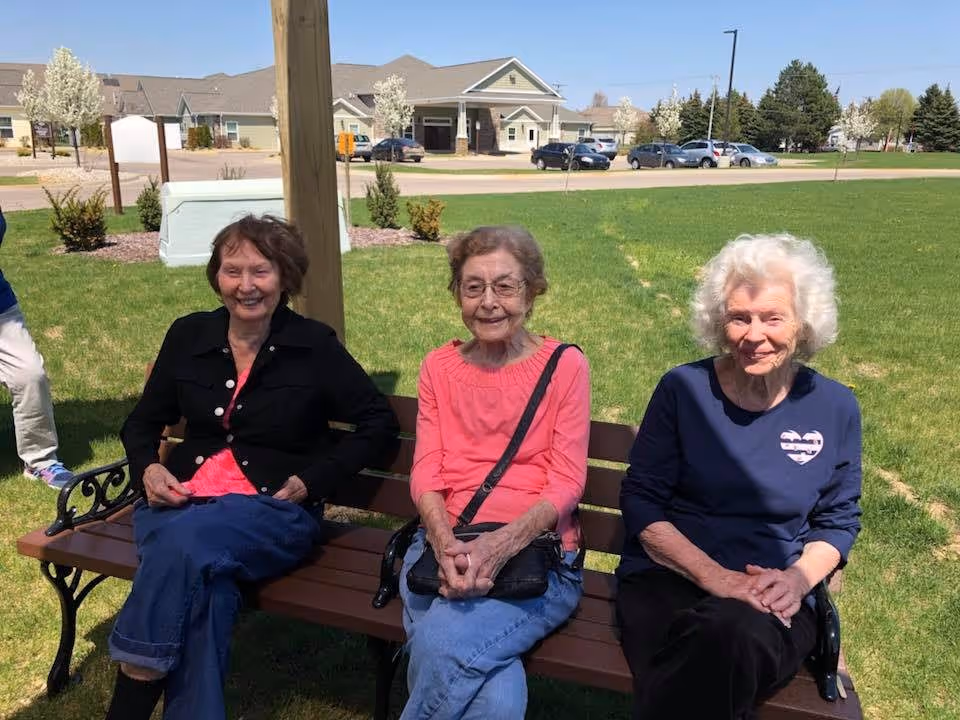 Three elderly women sitting on a wooden bench outdoors on a sunny day with green grass and a building in the background. They are smiling and appear to be enjoying the pleasant weather.
