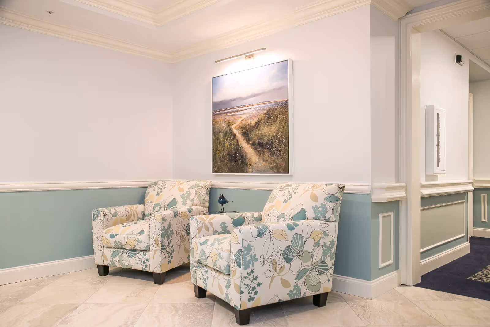 A cozy corner in a senior living facility with two floral-patterned armchairs placed against a light blue and white wall. Above the chairs is a framed painting of a sandy path through tall grass leading to a beach under a cloudy sky. The floor is tiled, and there is a hallway visible to the right.