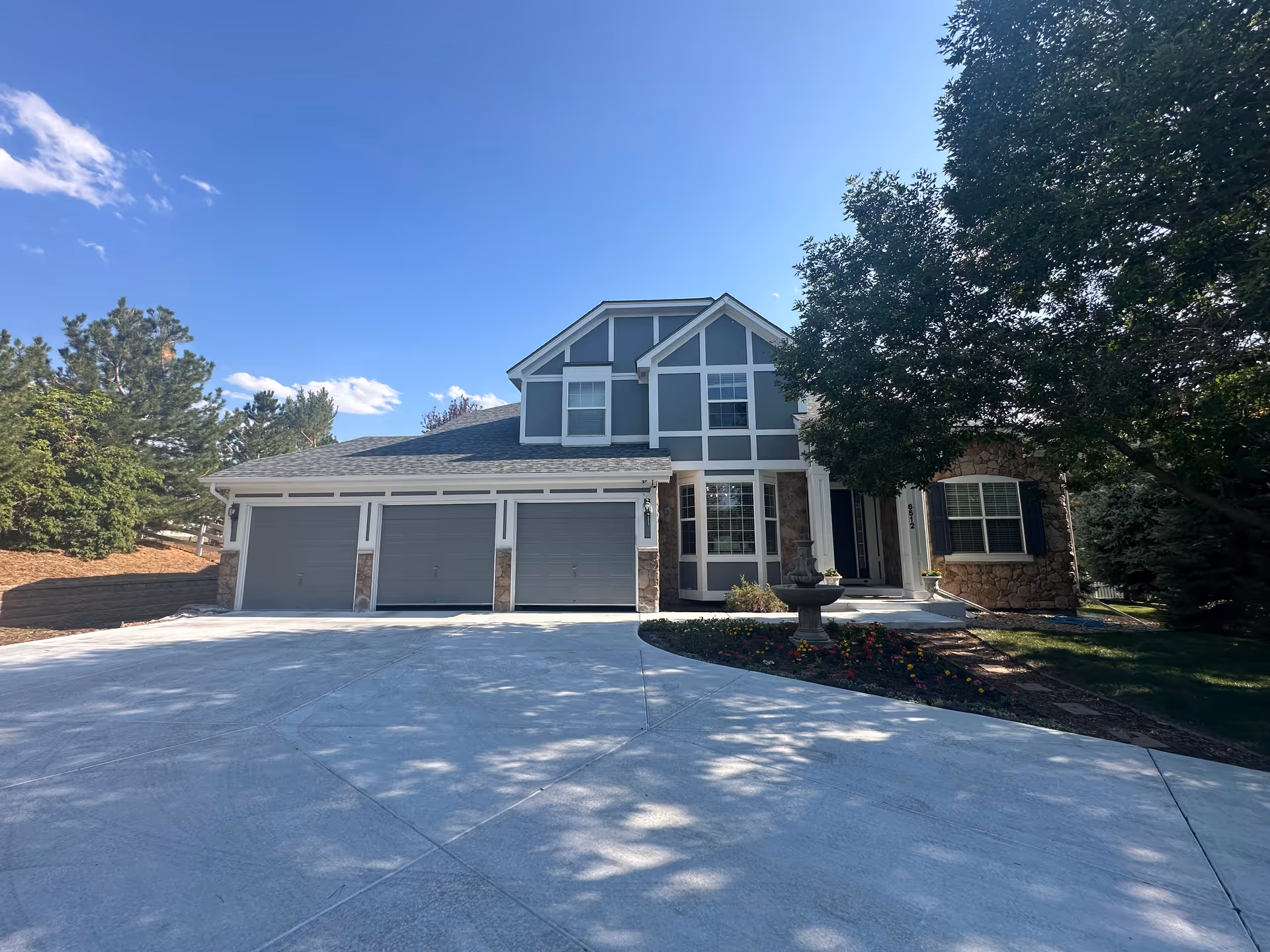 Front exterior view of a two-story house with a three-car garage, a concrete driveway, and a landscaped garden with a tree and a small fountain in front.
