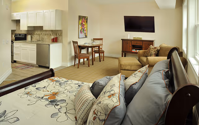 Interior view of a senior living facility room featuring a bed with floral bedding and multiple pillows in the foreground, a small dining table with two chairs, a kitchenette with white cabinets and appliances, and a living area with a sofa, ottoman, wooden TV stand, and a wall-mounted flat-screen TV. A colorful painting hangs on the wall near the dining area.