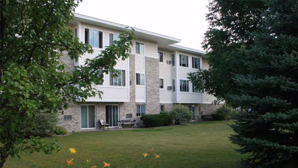 Three-story apartment building with a grassy lawn, patio chairs, and surrounding trees.