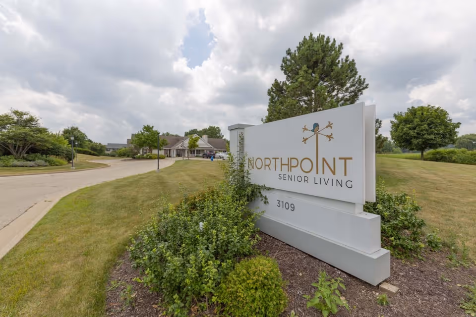 Outdoor view of the entrance to North Point Senior Living facility with a large white sign displaying the facility name and address 3109, surrounded by greenery and a driveway leading to the building under a cloudy sky.