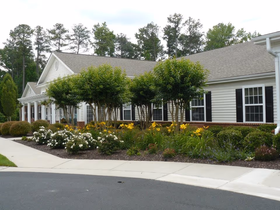 Exterior view of a single-story building with white siding and black shutters, surrounded by neatly landscaped bushes, small trees, and flowering plants. A paved driveway and sidewalk are visible in the foreground, with tall trees in the background.