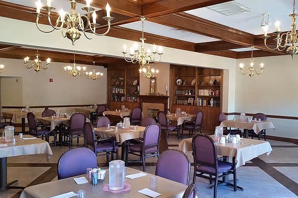 Dining room with multiple tables set for meals, purple chairs, chandeliers, and wooden built-in shelving.