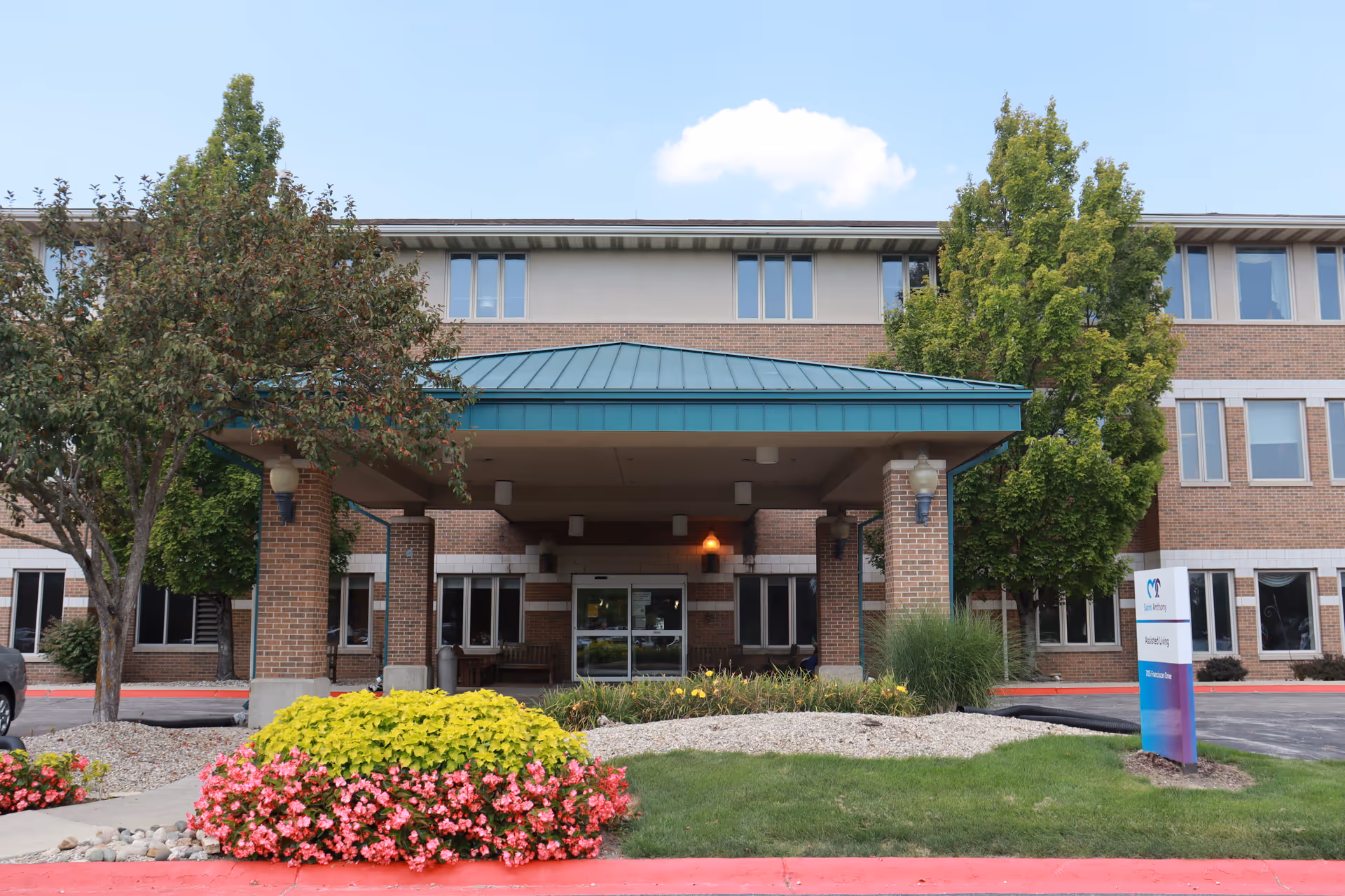 Front entrance of a three-story brick building with a teal metal roof over the entryway. There are trees and colorful flower beds in front, along with a sign that reads 'Saint Anthony, Assisted Living'. The sky is clear with a few clouds.