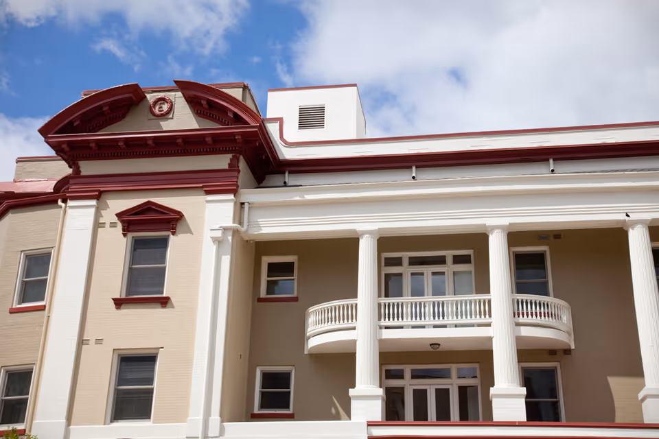 Exterior view of a beige and white building with red trim, featuring large white columns and a balcony under a partly cloudy blue sky.