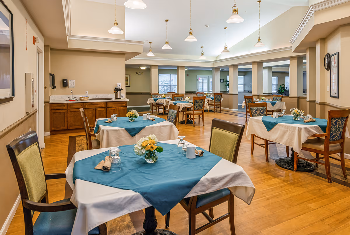 A bright communal dining room with tables covered in teal and white tablecloths, chairs, and floral centerpieces.