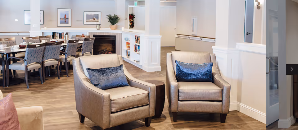 Two upholstered armchairs with blue pillows in a bright common lounge area with a dining table, fireplace, and bookshelves.