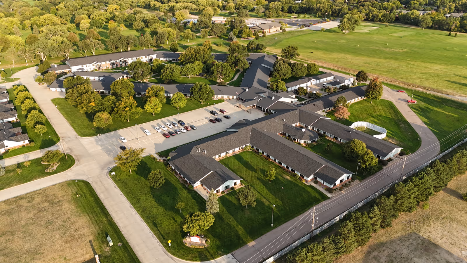Aerial view of Riverside Lodge Retirement Community showing multiple single-story buildings with dark roofs arranged around green lawns and trees. There is a parking lot with several cars, surrounding roads, and open grassy areas including a golf course in the background.