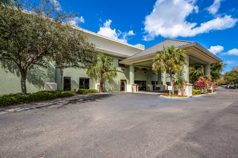 Front entrance of a senior living facility with a covered porte-cochère, palm trees, and a paved driveway under a blue sky.