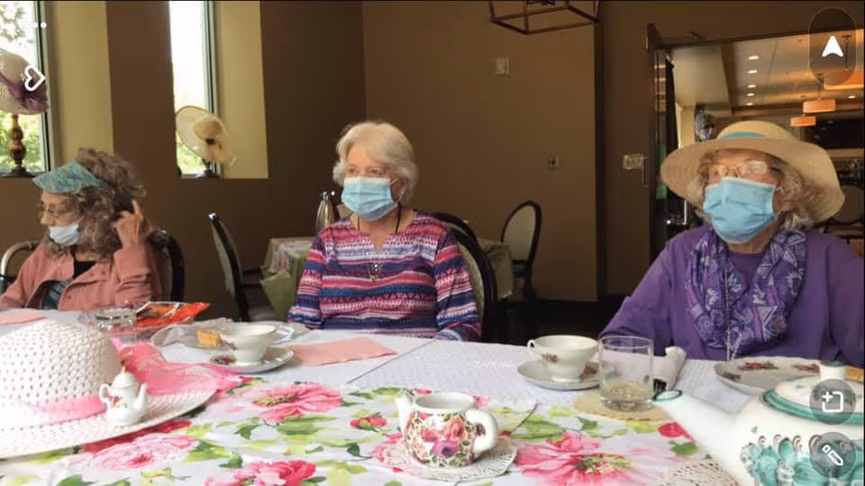 Three elderly women wearing face masks sitting around a table with floral tablecloth and tea set in a dining area. Two women are wearing hats, and the room has warm lighting with windows in the background.