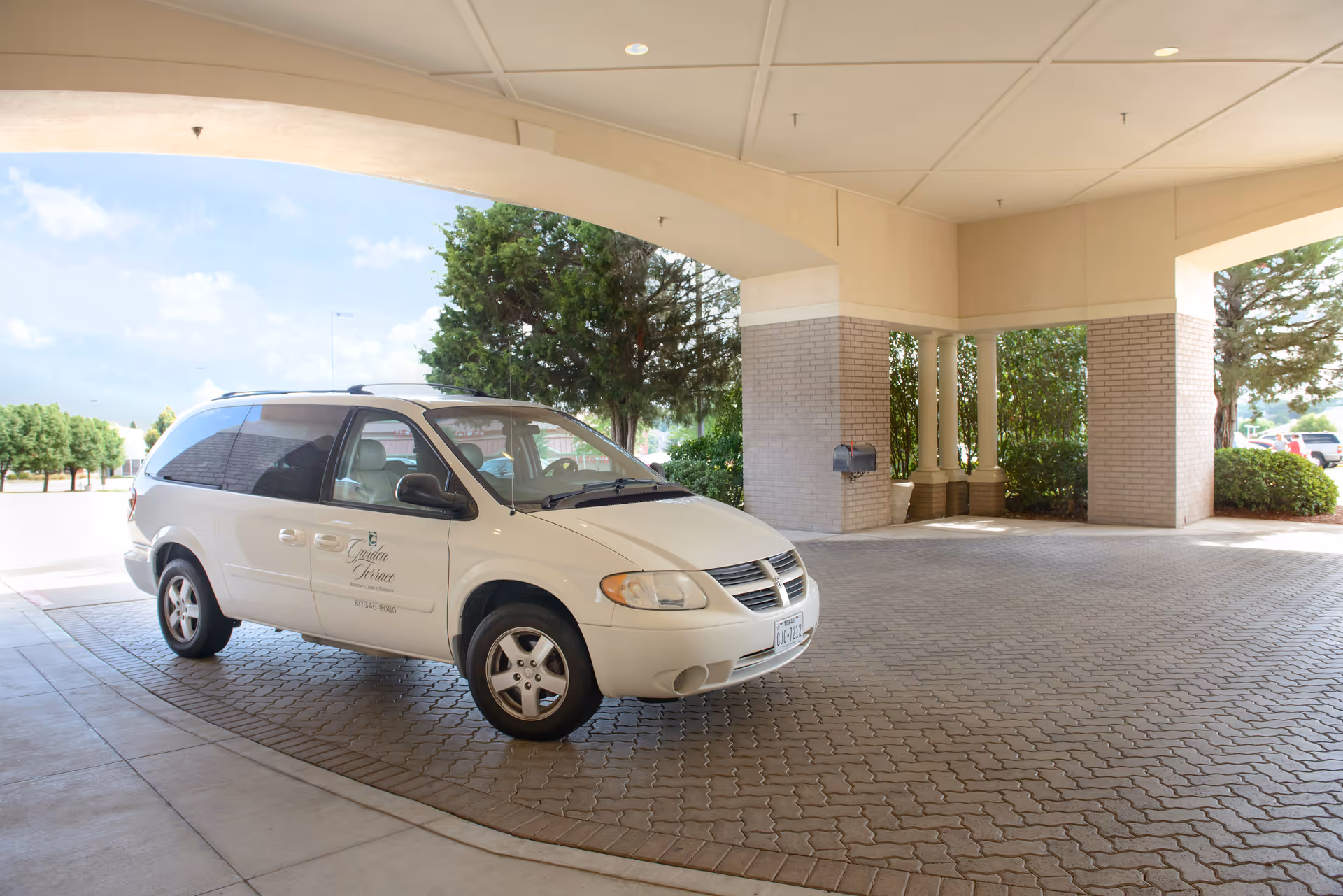 A white minivan with the logo and contact information of Garden Terrace at Fort Worth parked under a covered driveway area with brick pillars and greenery in the background.