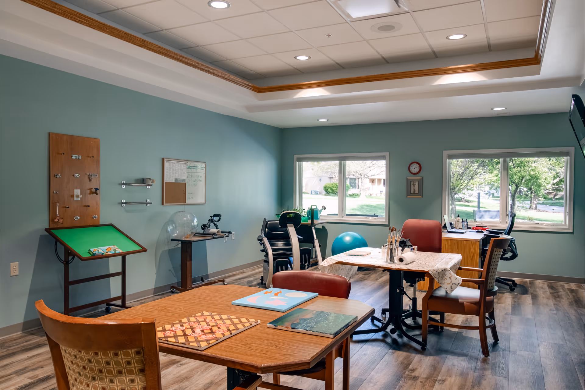 Bright communal activity room with tables and chairs, exercise equipment, and large windows overlooking trees.