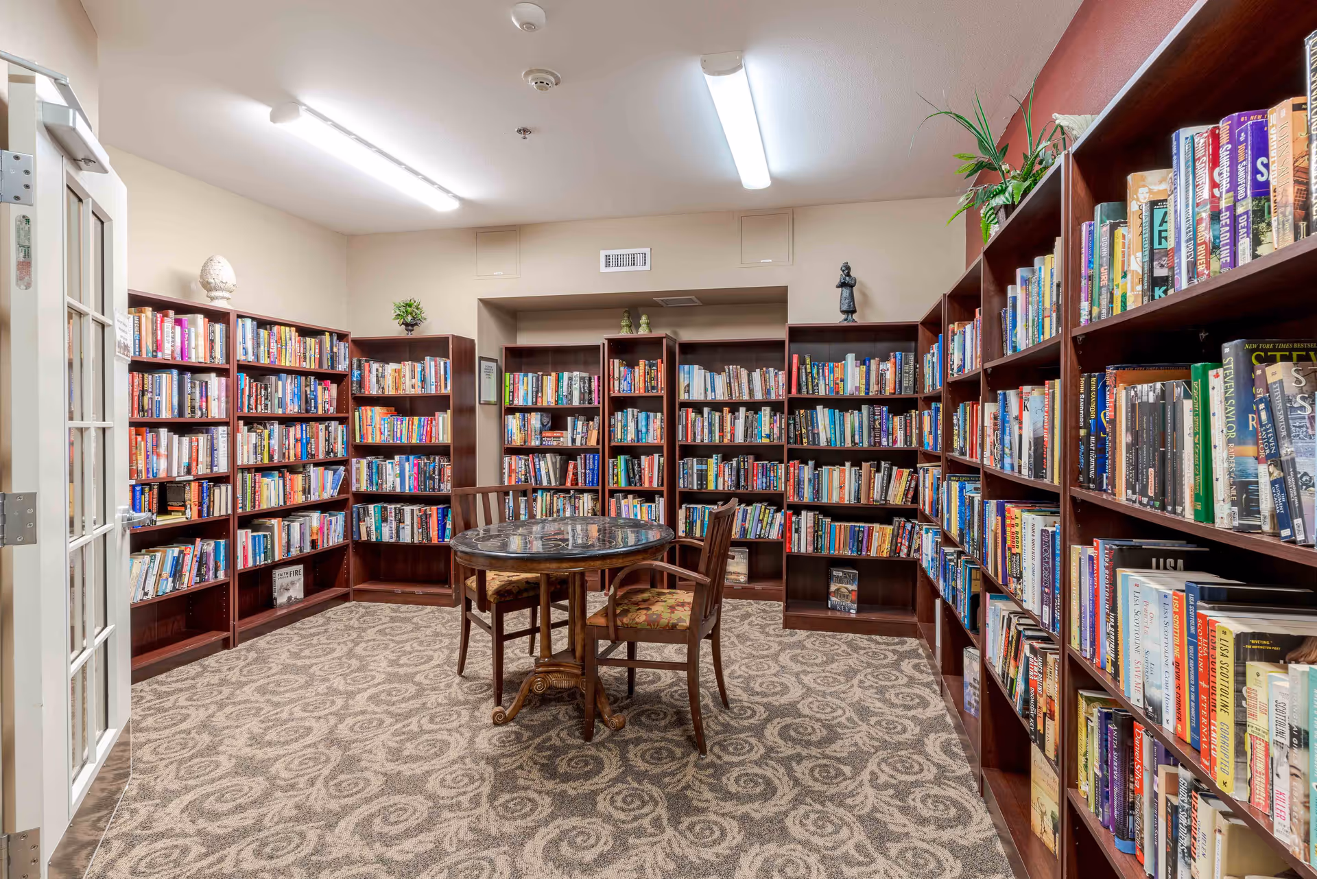 A cozy library room with multiple wooden bookshelves filled with books lining the walls. In the center, there is a round table with a decorative top and three wooden chairs with patterned cushions. The room has patterned carpet flooring and is well-lit with ceiling lights. There are small decorative plants and figurines on top of the bookshelves.