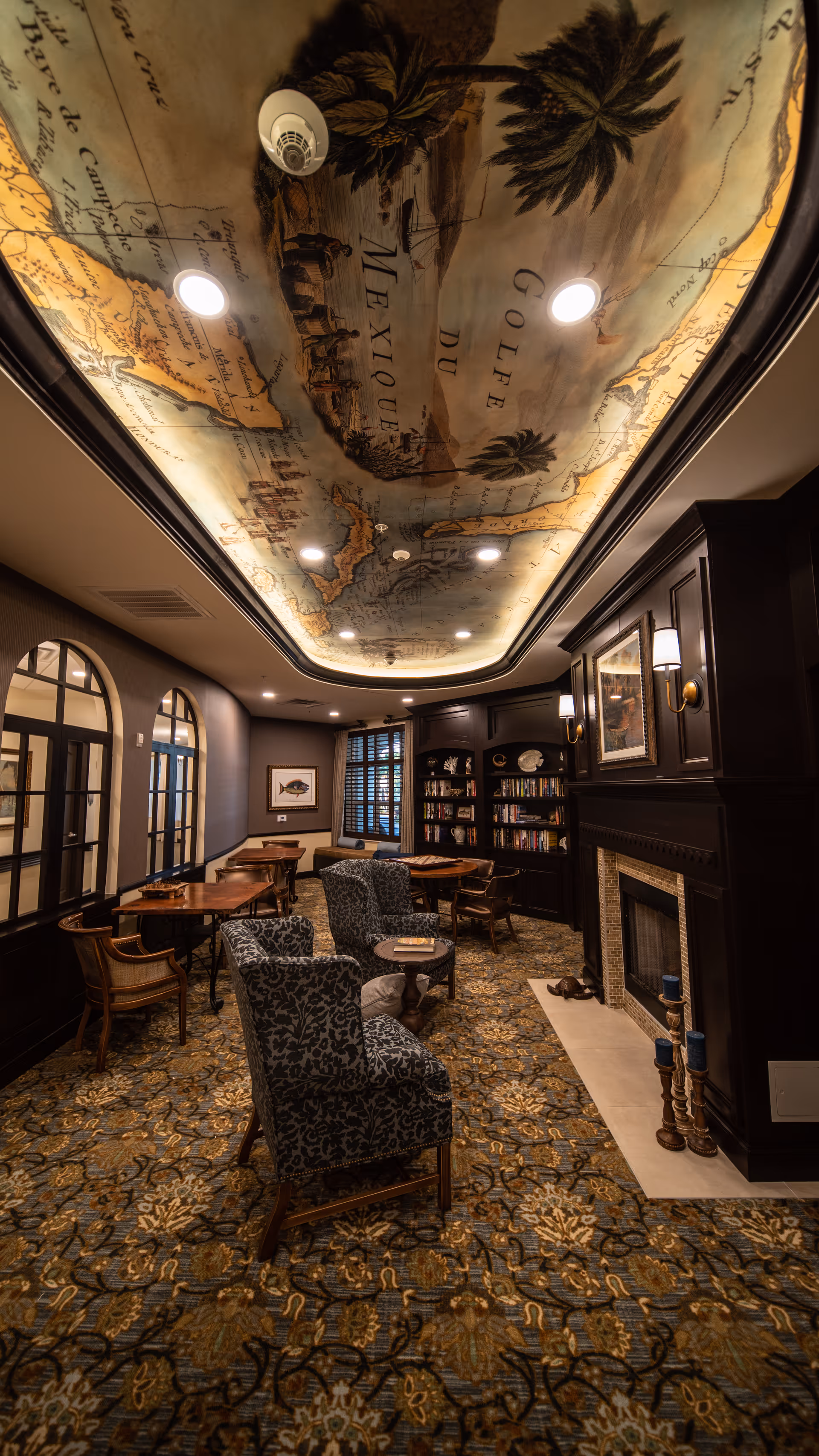A cozy, elegant common room with patterned carpet, upholstered armchairs, wooden tables and chairs, a fireplace with candles, built-in bookshelves filled with books, framed artwork on the walls, and a ceiling featuring a large illuminated vintage map design.