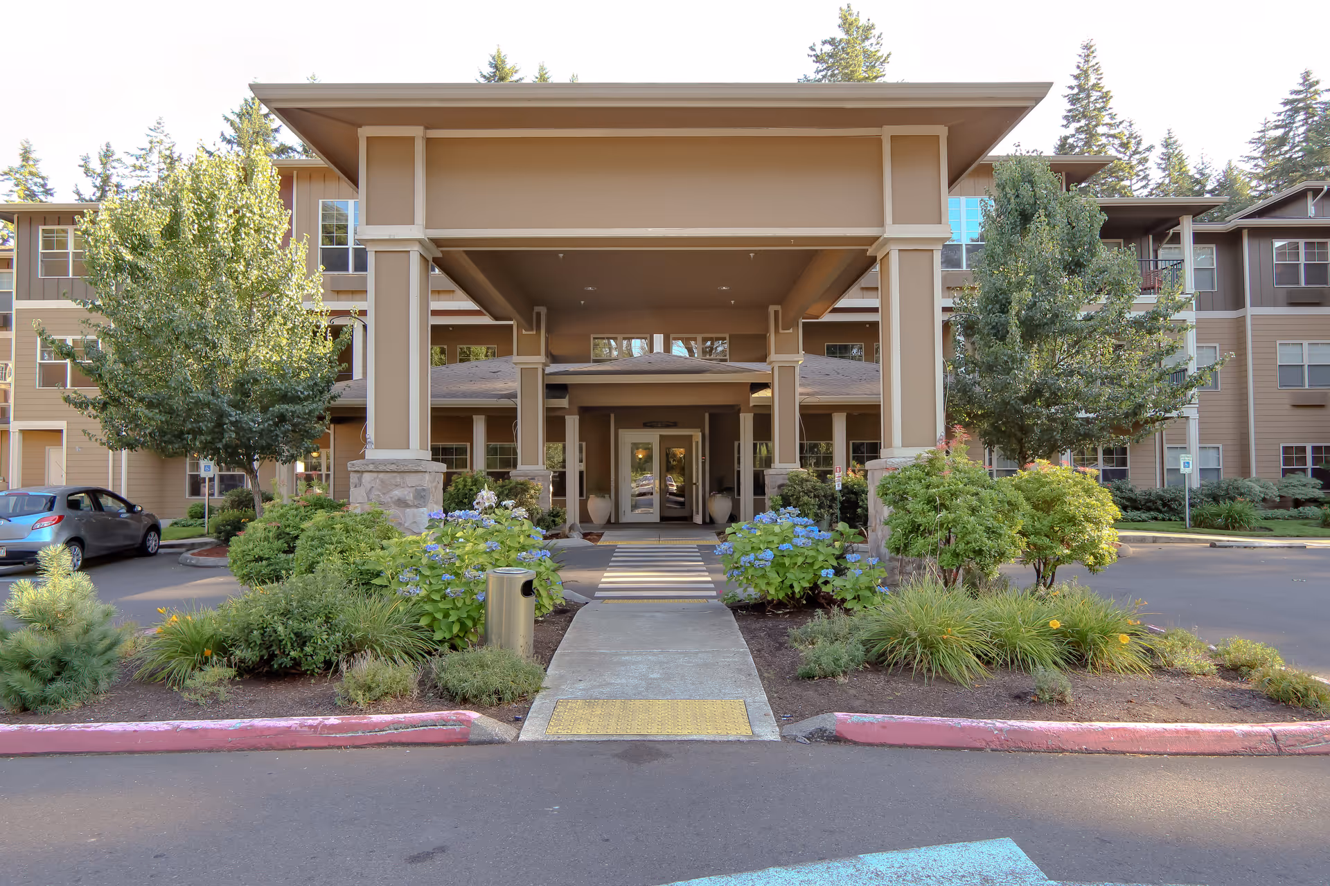 Front entrance of a senior living facility with a covered drop-off area, surrounded by landscaped greenery and trees, with a multi-story building in the background.