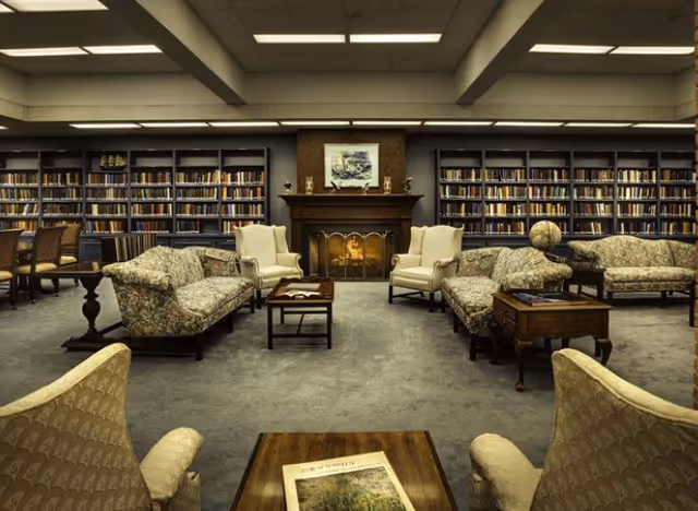 A cozy library room with multiple bookshelves filled with books along the back wall. The room features a fireplace in the center with a painting above it. There are several upholstered armchairs and sofas arranged around small wooden tables, creating a comfortable seating area. The lighting is provided by ceiling lights, and the carpet is a muted gray color.