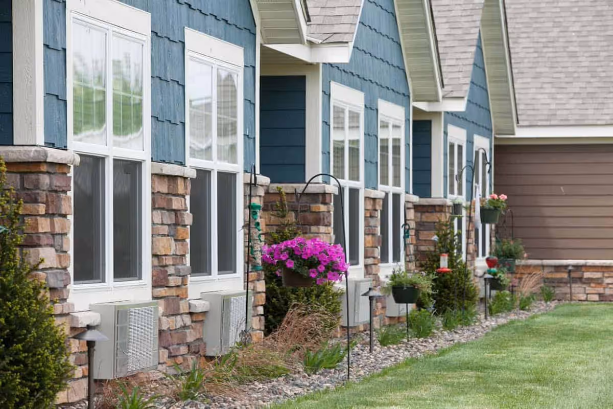 Exterior side view of a senior living facility building with blue siding, stone accents, multiple white-framed windows, hanging flower pots with pink flowers, and a well-maintained lawn and garden area.