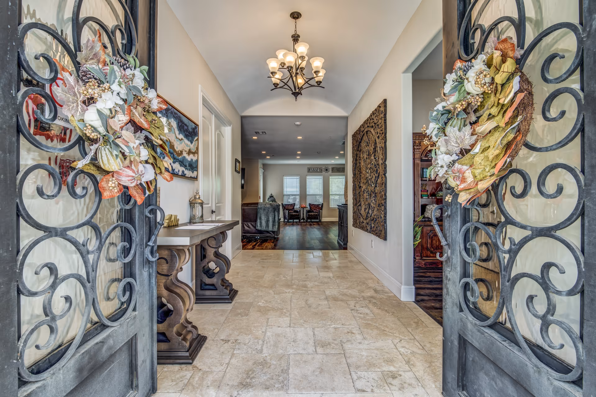 View through decorative wrought iron double doors with autumn-themed wreaths into a hallway with beige tiled floor, a chandelier hanging from a vaulted ceiling, a console table on the left, and a large carved wooden wall art on the right. The hallway leads to a living area with chairs and windows in the background.