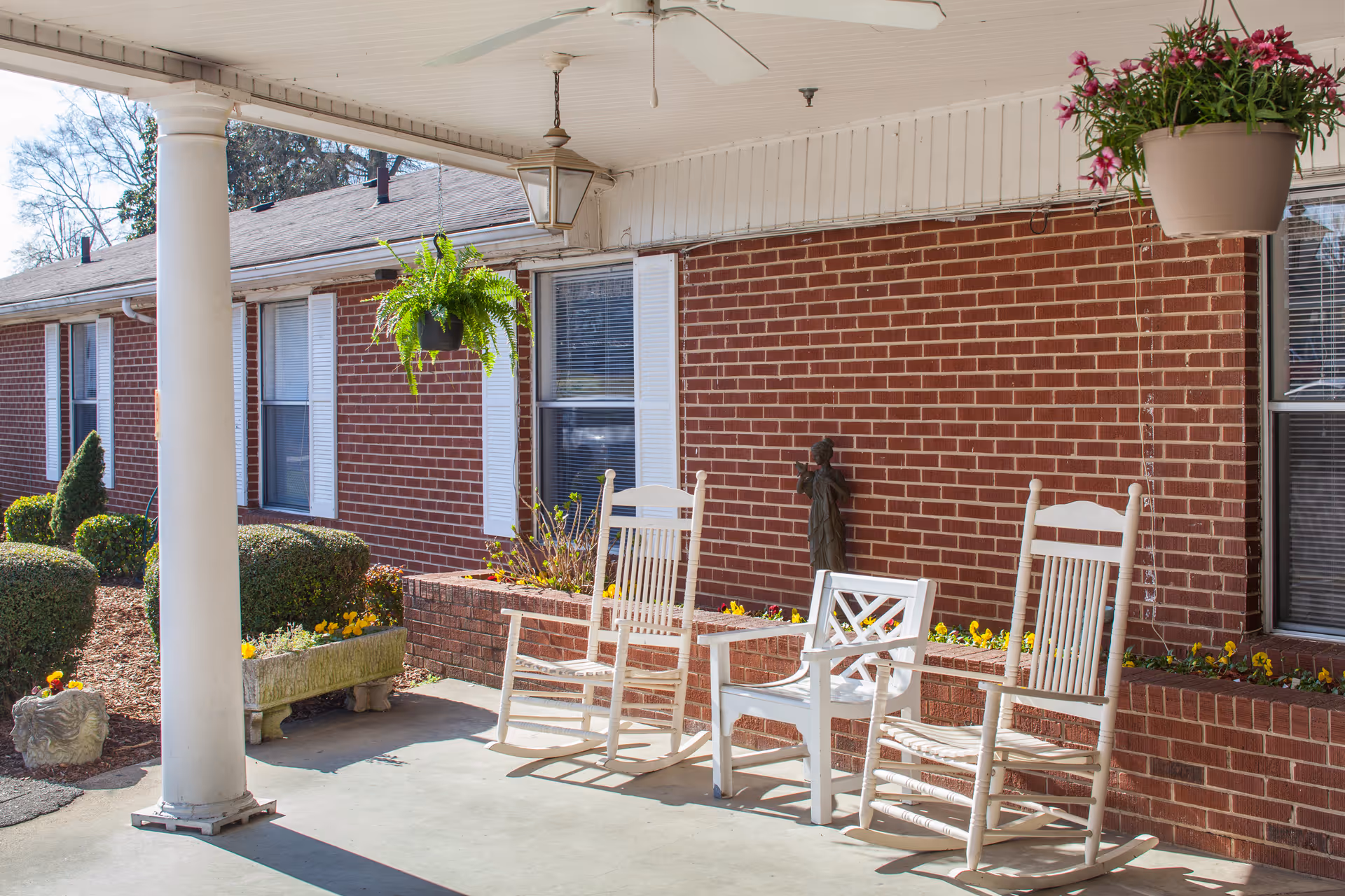 Covered brick porch with white rocking chairs, hanging plants, and decorative columns.