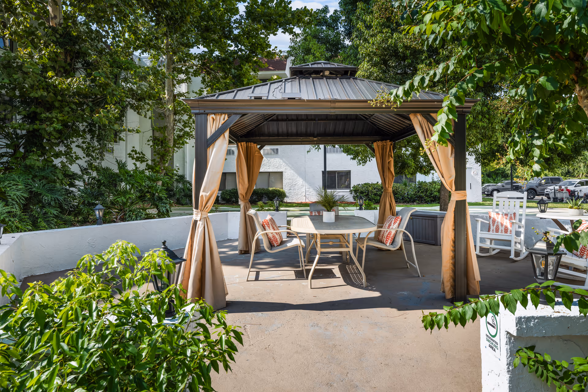 Outdoor patio area with a metal gazebo featuring beige curtains tied to the posts. Under the gazebo is a table with four chairs, each with a red and white patterned cushion. Surrounding the patio are green trees and bushes, with a white building and parked cars visible in the background.